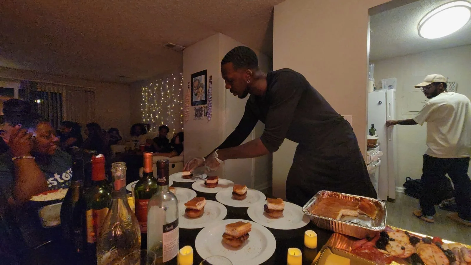 Person serving food at a dinner party with guests seated around a table, bottles of alcohol, and decorative string lights in the background.