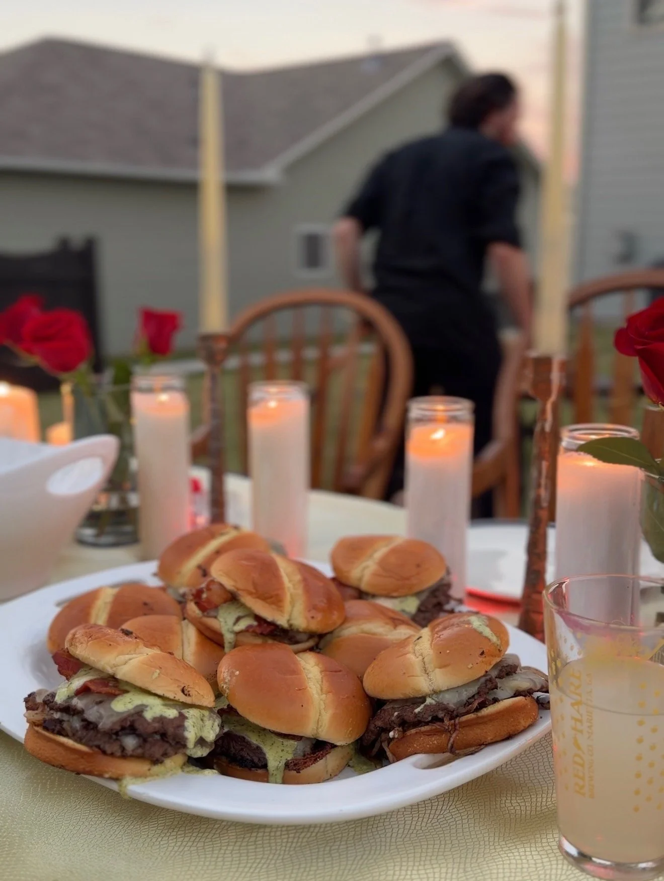 Plate of mini sandwiches on a table with lit candles, flowers, and a drink, outdoor setting with a person in the background.
