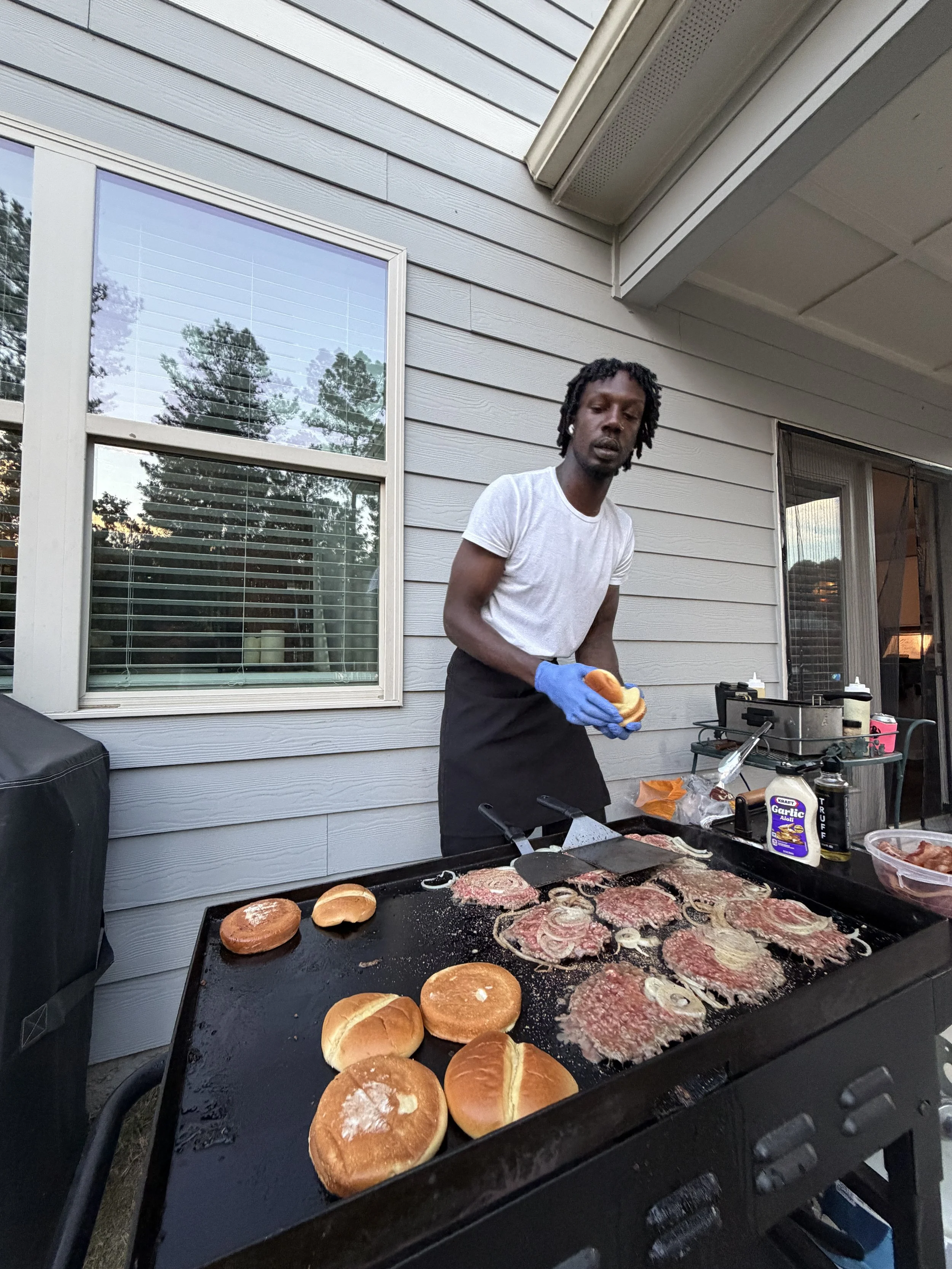 Man cooking meat and buns on an outdoor grill, wearing a white t-shirt, black apron, and blue gloves, with house siding and a window in the background.