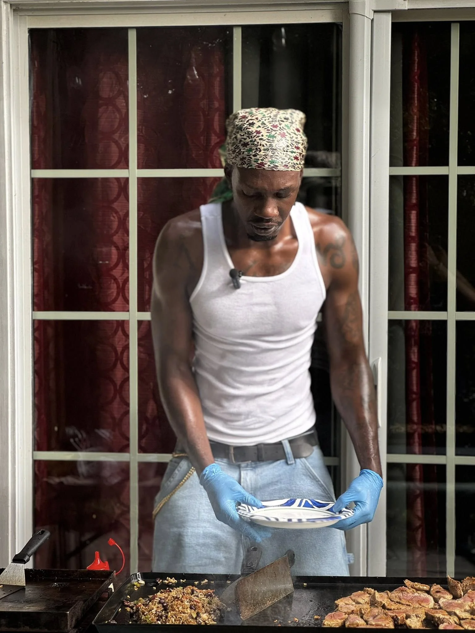A man wearing a white tank top, a colorful bandana, and blue gloves stands behind a grill, holding a patterned plate with food, with cooked meat and a side dish cooking on the grill.