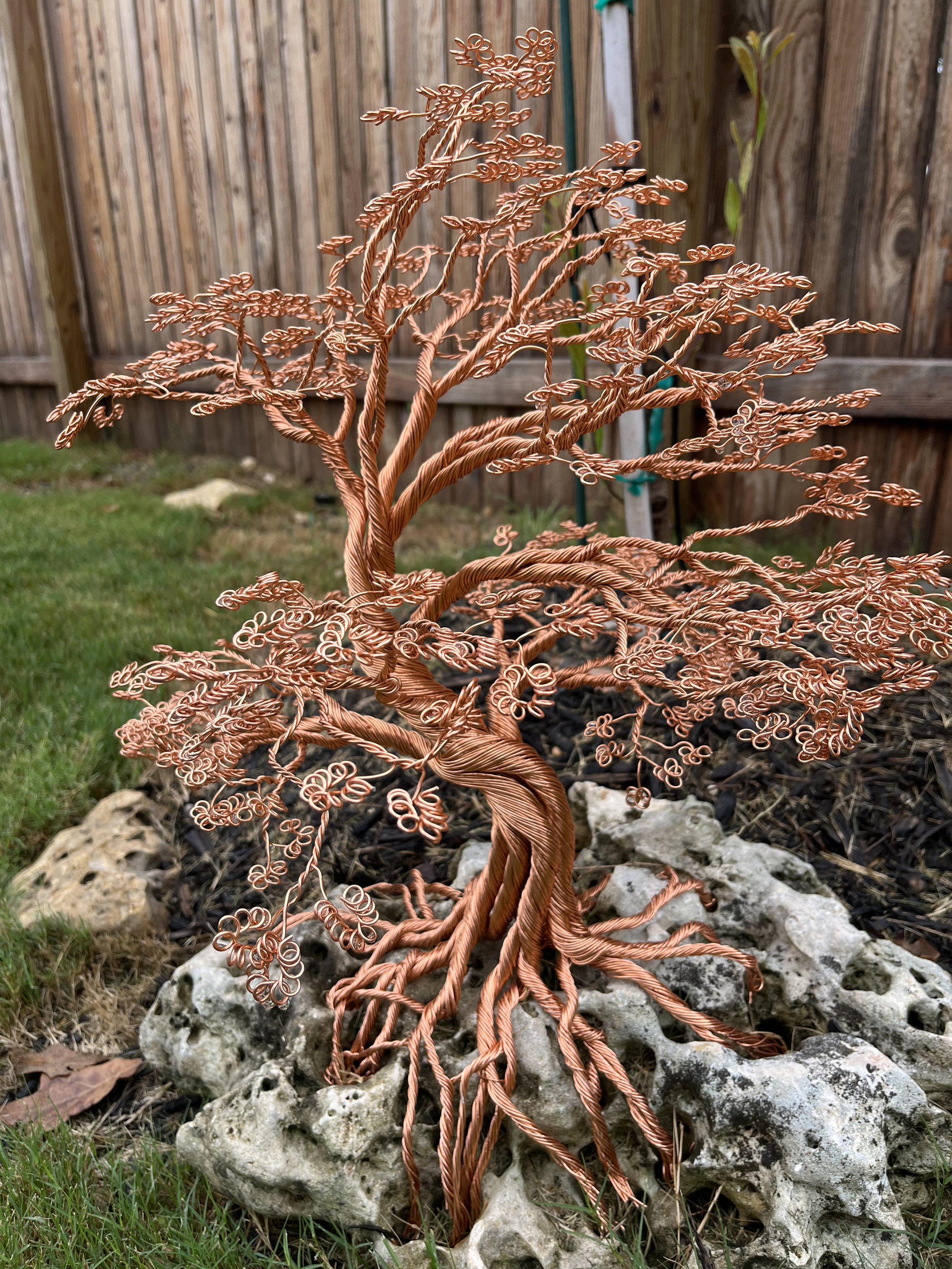 An intricate sculpture resembling a tree made of twisted and curled copper wire, mounted on rocks in a garden with a wooden fence in the background.
