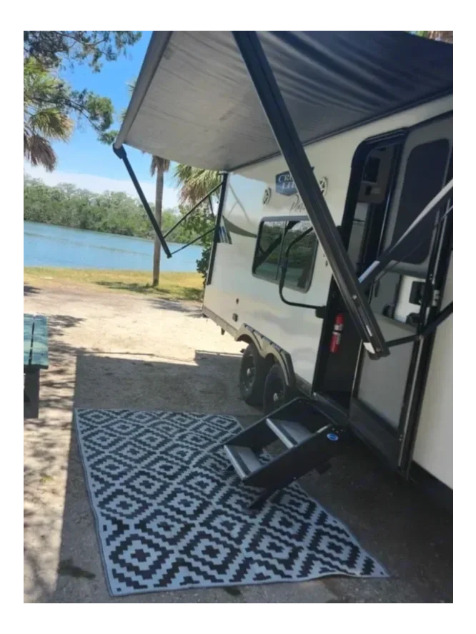 A camper parked outdoors near a lake, with an extended awning and a black and white geometric rug on the ground.