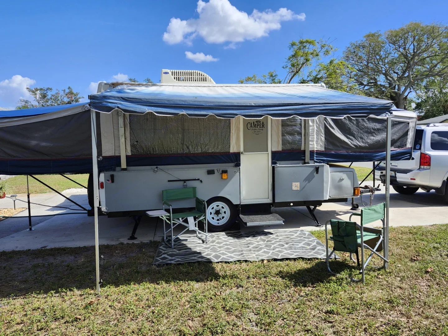 A pop-up camper trailer set up on a grassy area with two green camping chairs and a gray patterned outdoor rug underneath. The camper is white with a blue and gray awning extended, and there are trees and a blue sky in the background.