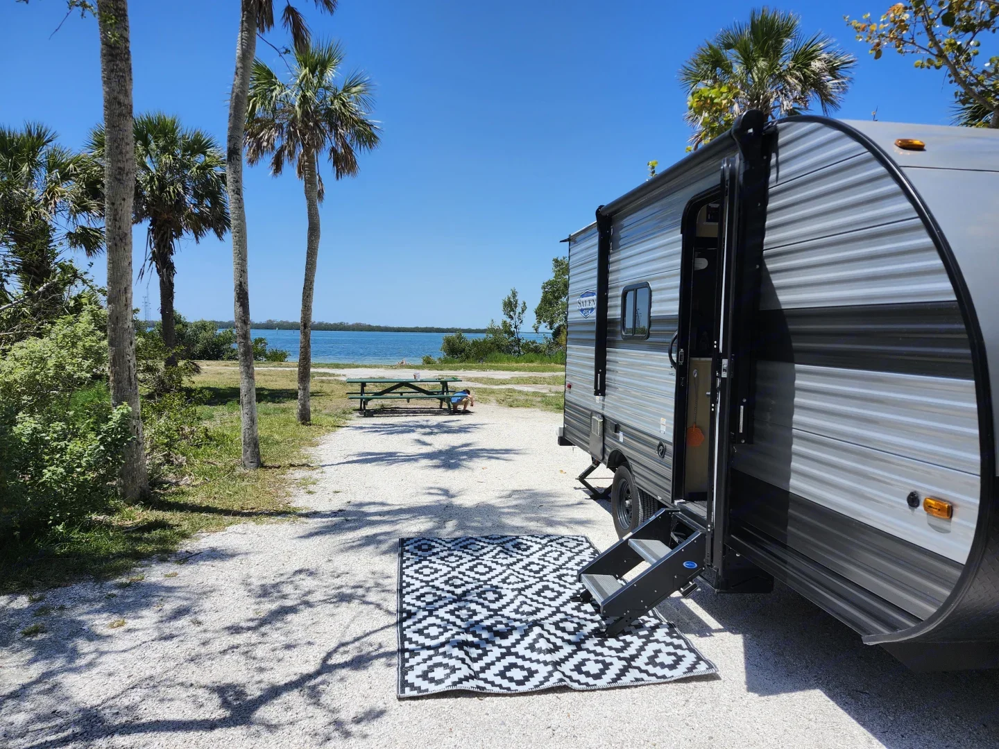 A camping trailer parked near a lakeside with palm trees, a picnic table, and a patterned outdoor rug on a sunny day.
