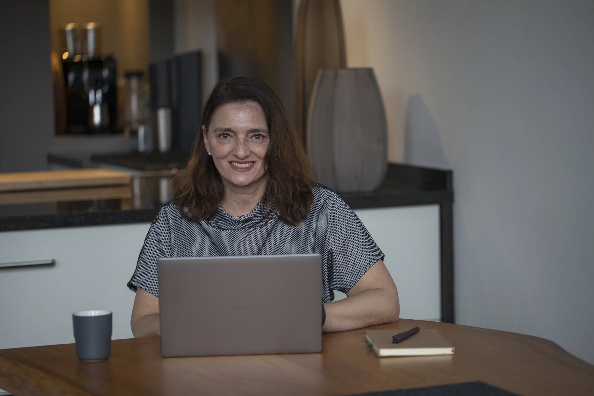 A woman with long brown hair smiling, sitting at a wooden table with a laptop, notebook, pen, and a cup.
