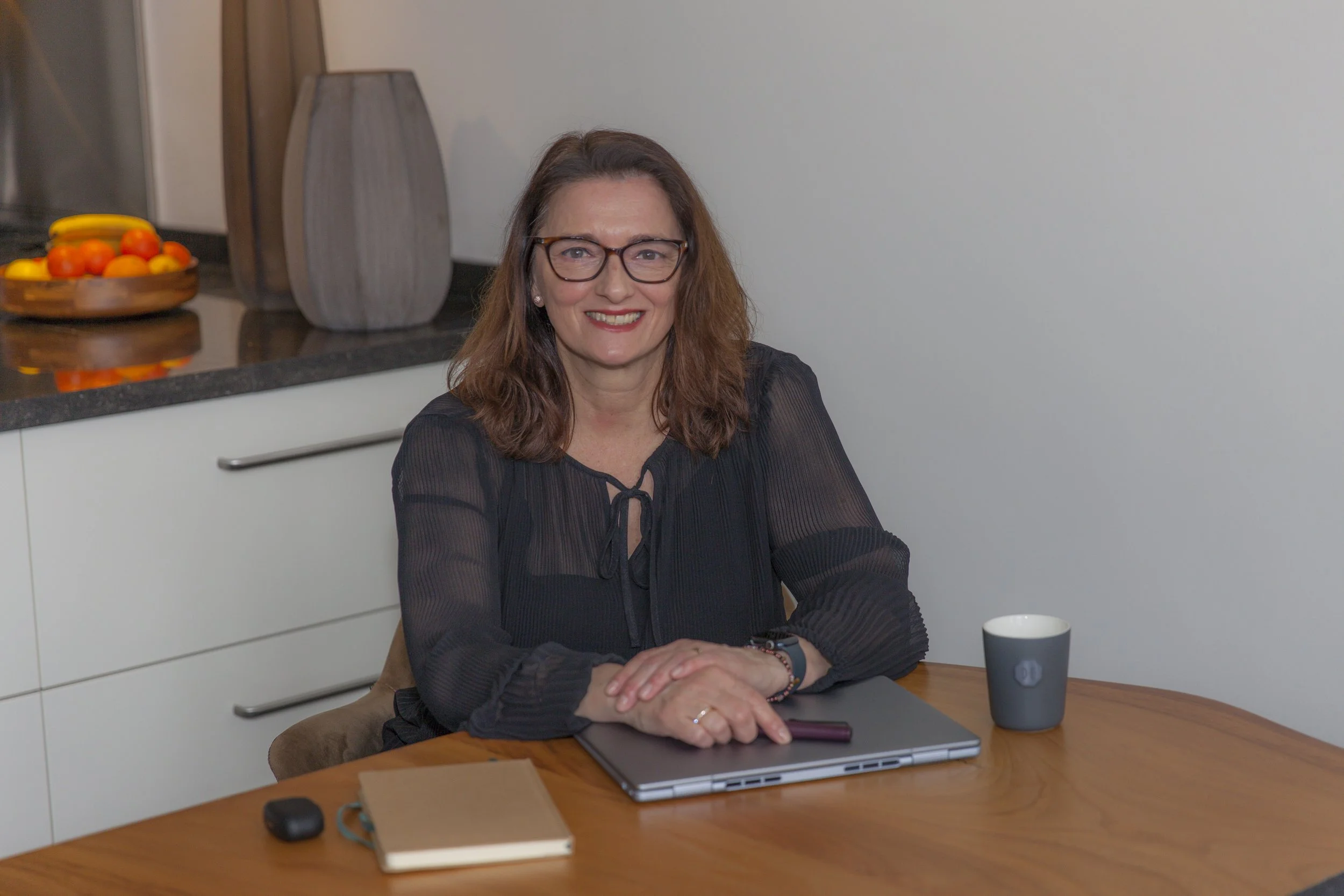 A woman with long brown hair, glasses, and a black blouse sitting at a wooden table with a laptop, a notebook, a phone, a mug, and a computer mouse. She is smiling and looking at the camera in a modern kitchen with white cabinets, a bowl of fruit, and decorative vases in the background.