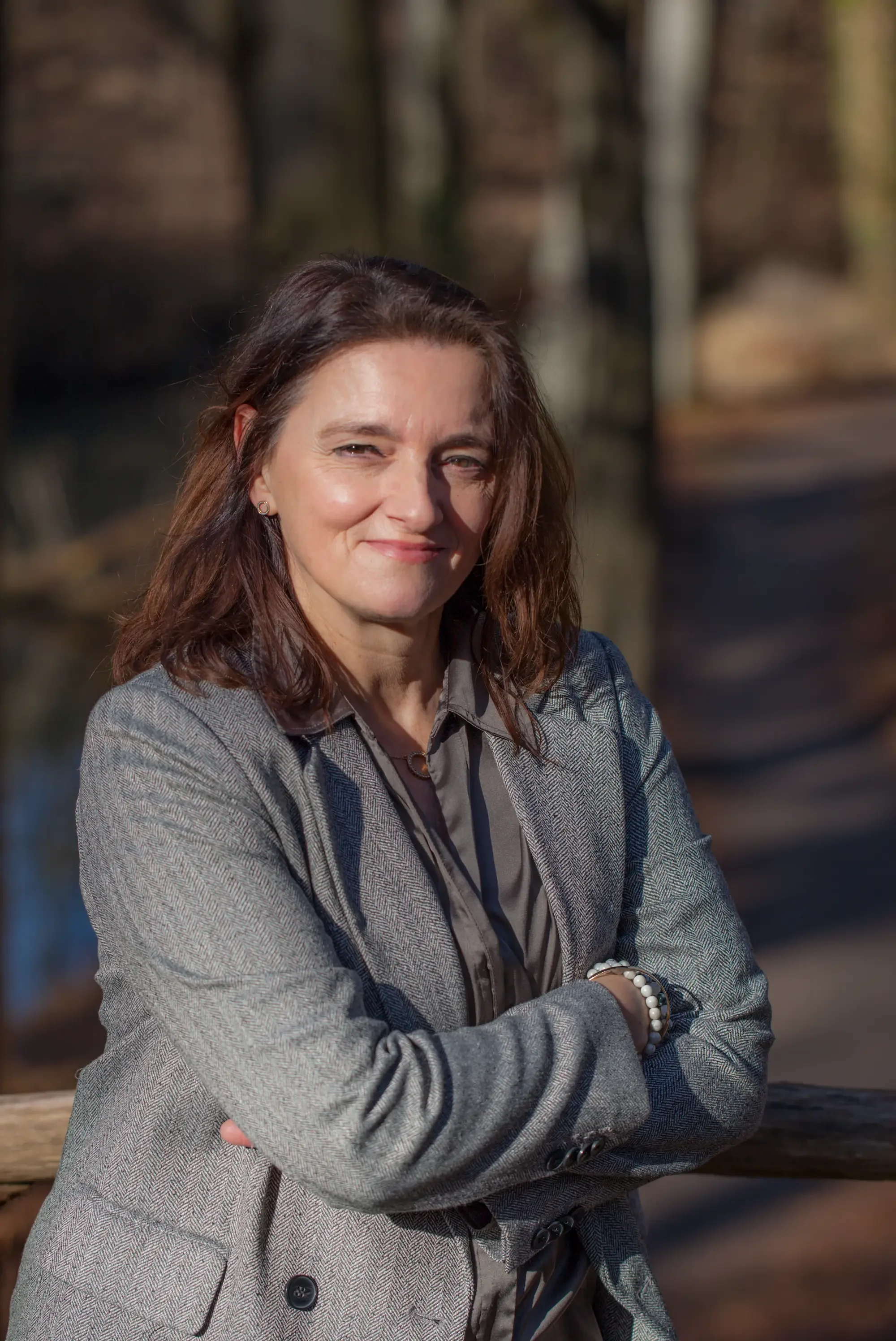 A woman with brown hair, dressed in a grey blazer and shirt, standing outdoors with her arms crossed, smiling slightly, with a blurred background of trees and natural surroundings.