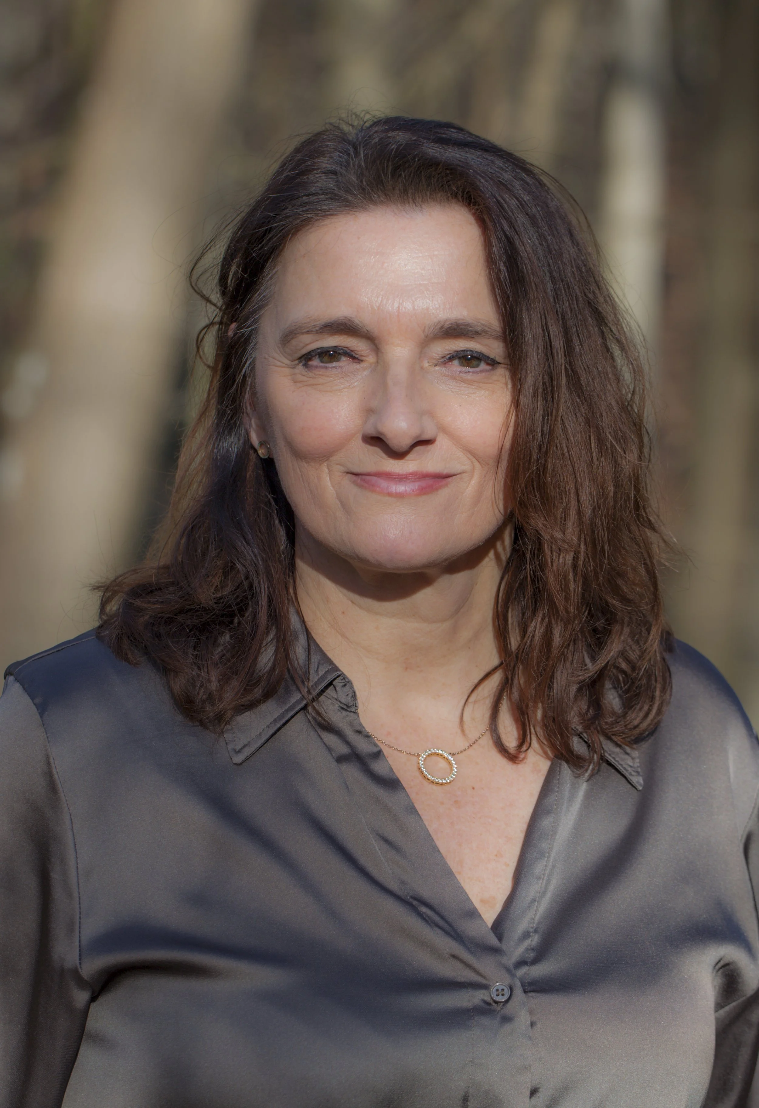 A woman with shoulder-length brown hair and light skin, wearing a gray button-up shirt and a necklace with a circular pendant, standing outdoors in a sunny setting with blurred trees in the background.