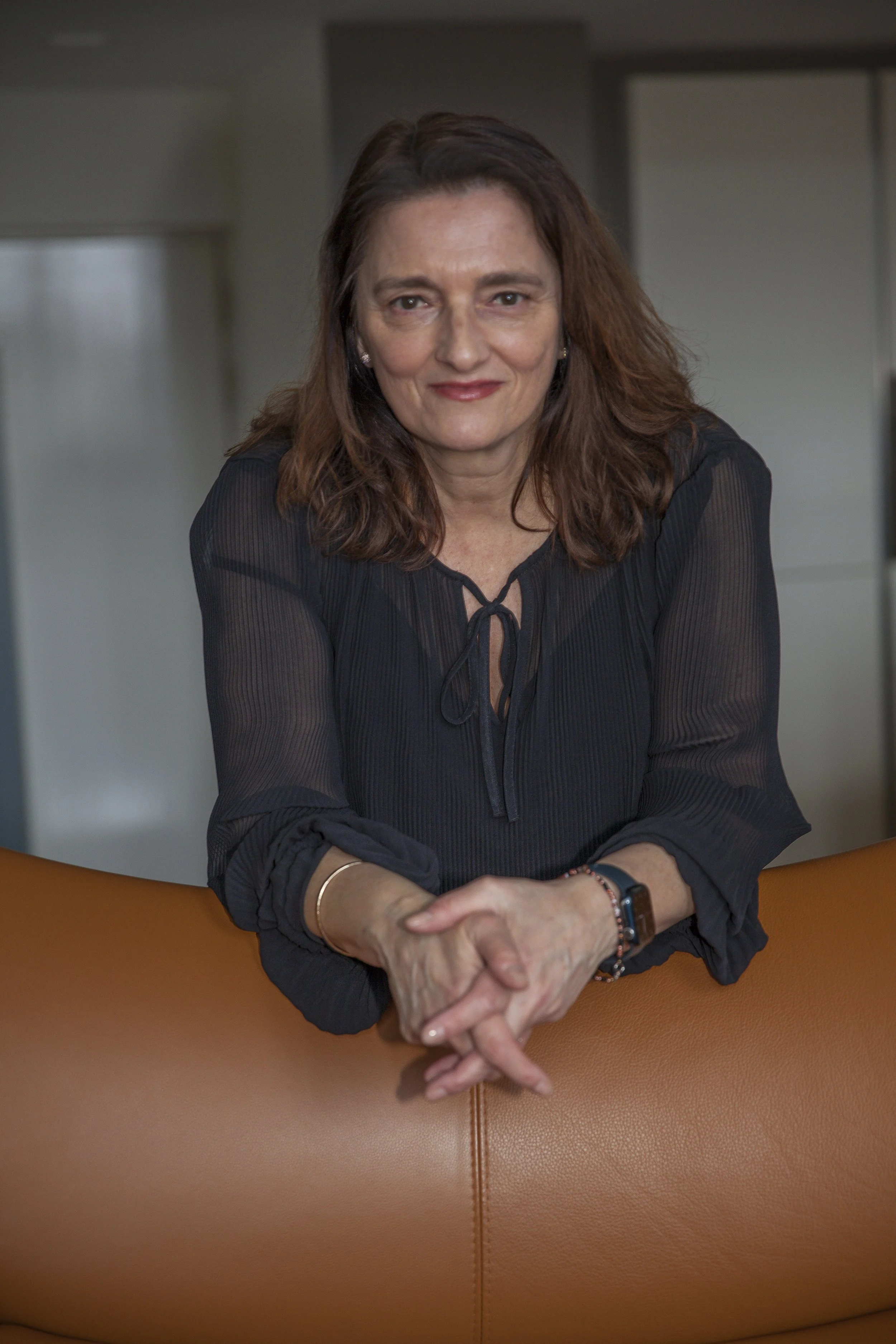 A woman with brown hair, wearing a black blouse, leaning on a tan leather surface, smiling at the camera.