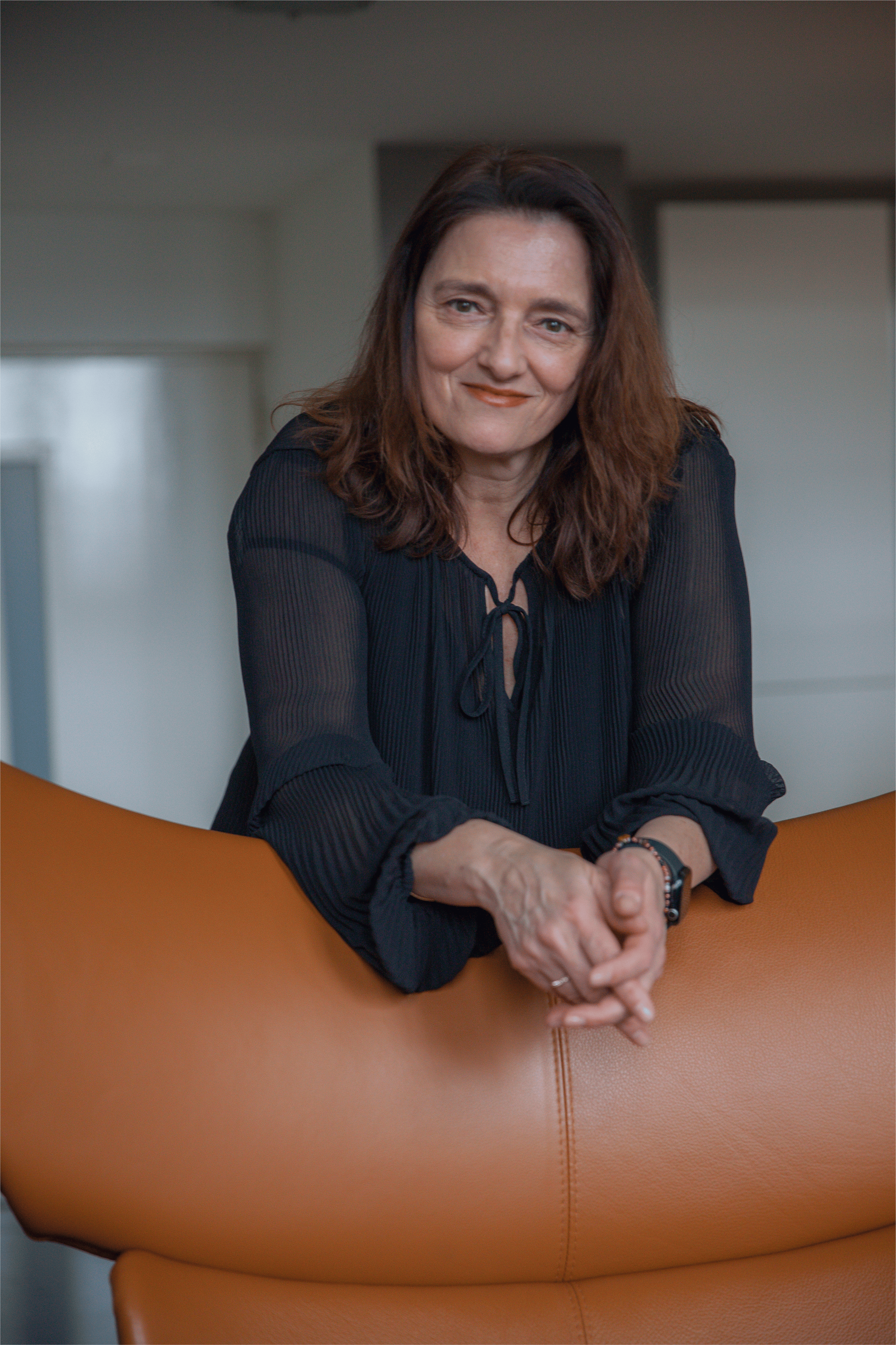 A woman with shoulder-length brown hair and light skin standing behind a brown fauteuil in a modern interior. She is smiling slightly and leaning forward, resting her arms on the chair, wearing a black blouse with a tie-front detail.