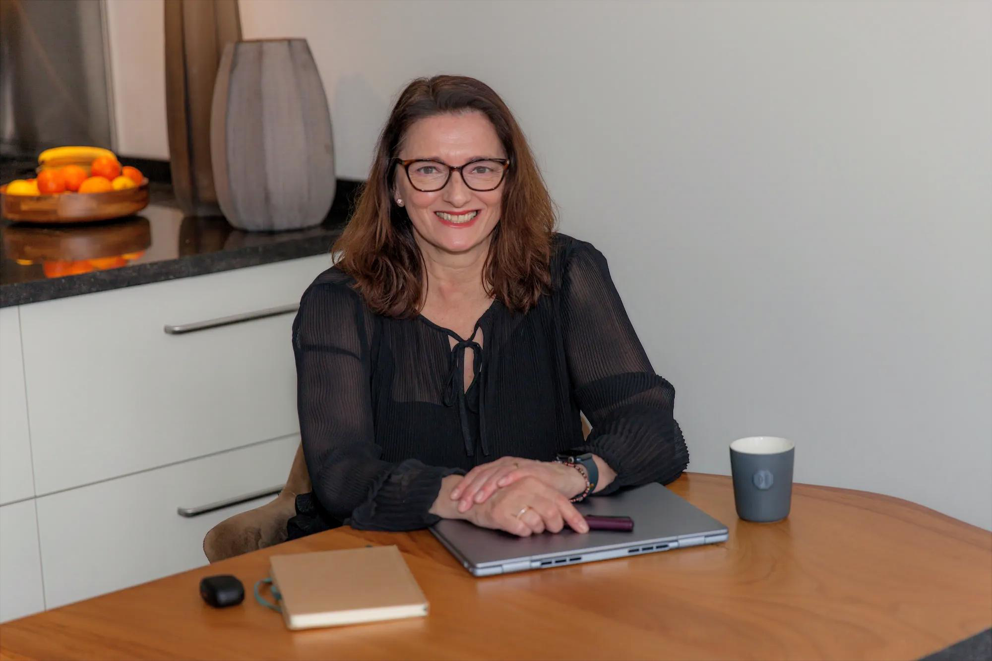 Woman with brown hair and glasses smiling while sitting at a wooden table with a laptop, notebook, and a mug.