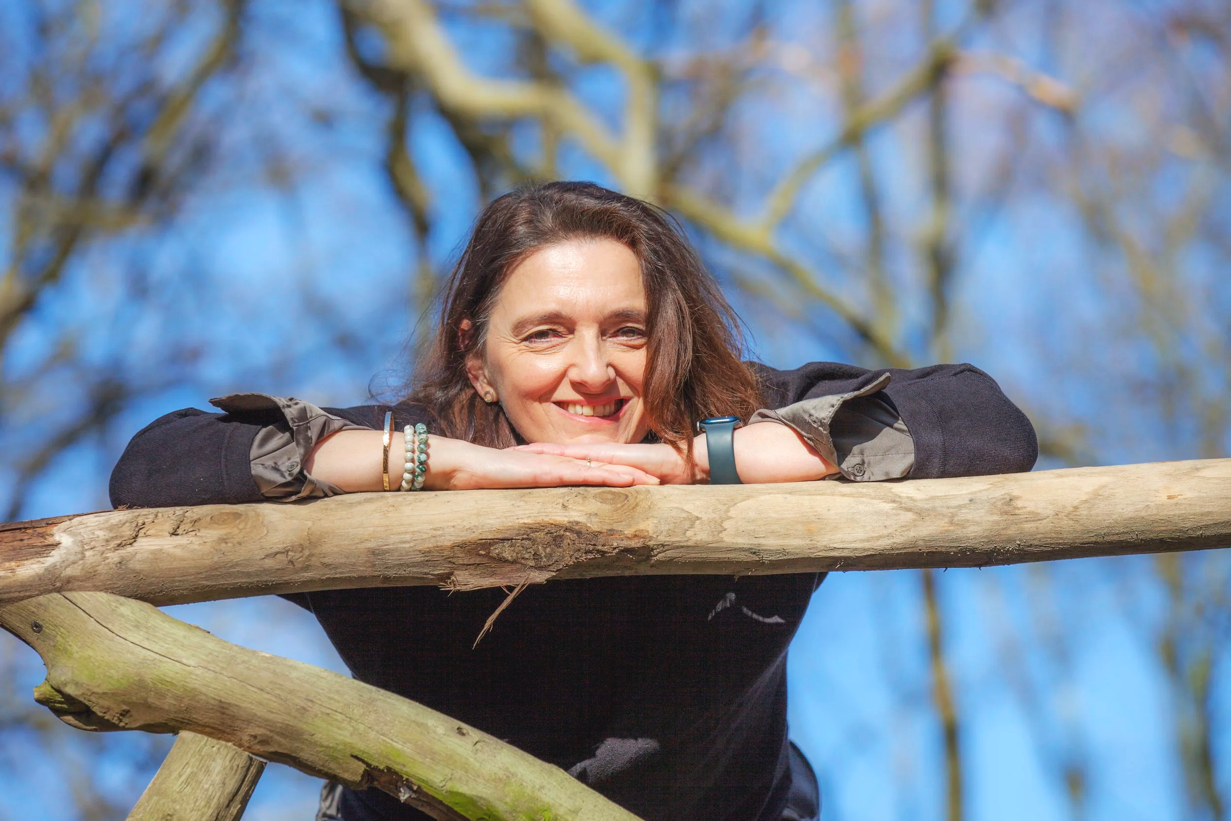 A woman resting her head on her arms on a wooden railing outdoors, smiling at the camera with trees and blue sky in the background.