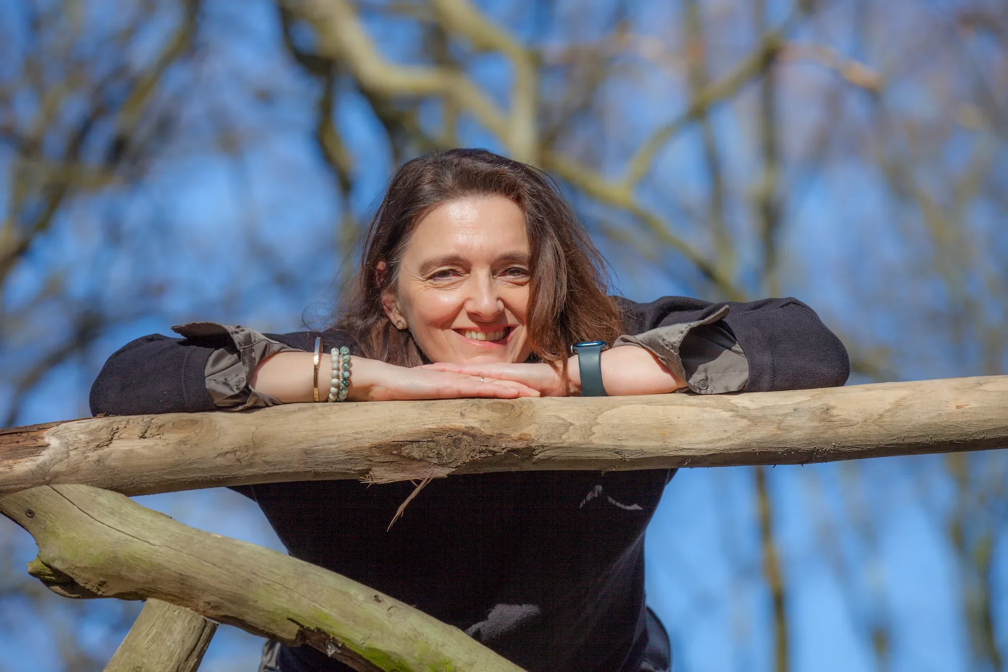 Woman with brown hair smiling and resting her chin on her arms, leaning on a wooden railing in an outdoor setting with a blue sky and tree branches in the background.