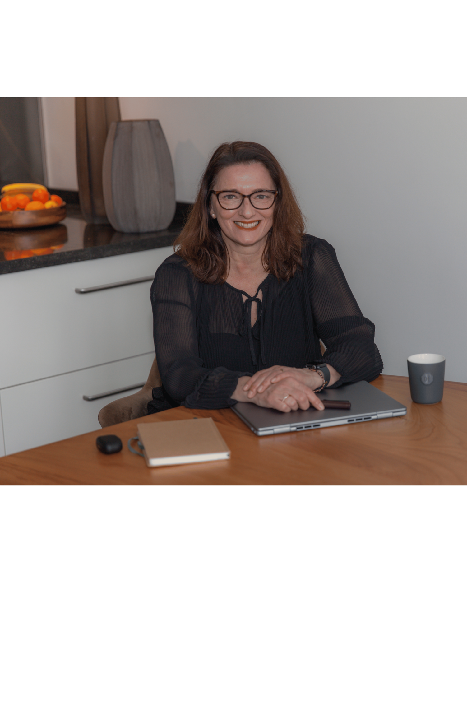 A woman with brown hair, glasses, and a black blouse sitting at a wooden table, smiling at the camera. The table has a laptop, a notebook, a computer mouse, a smartphone, and a coffee mug. In the background, there is a kitchen countertop with a bowl