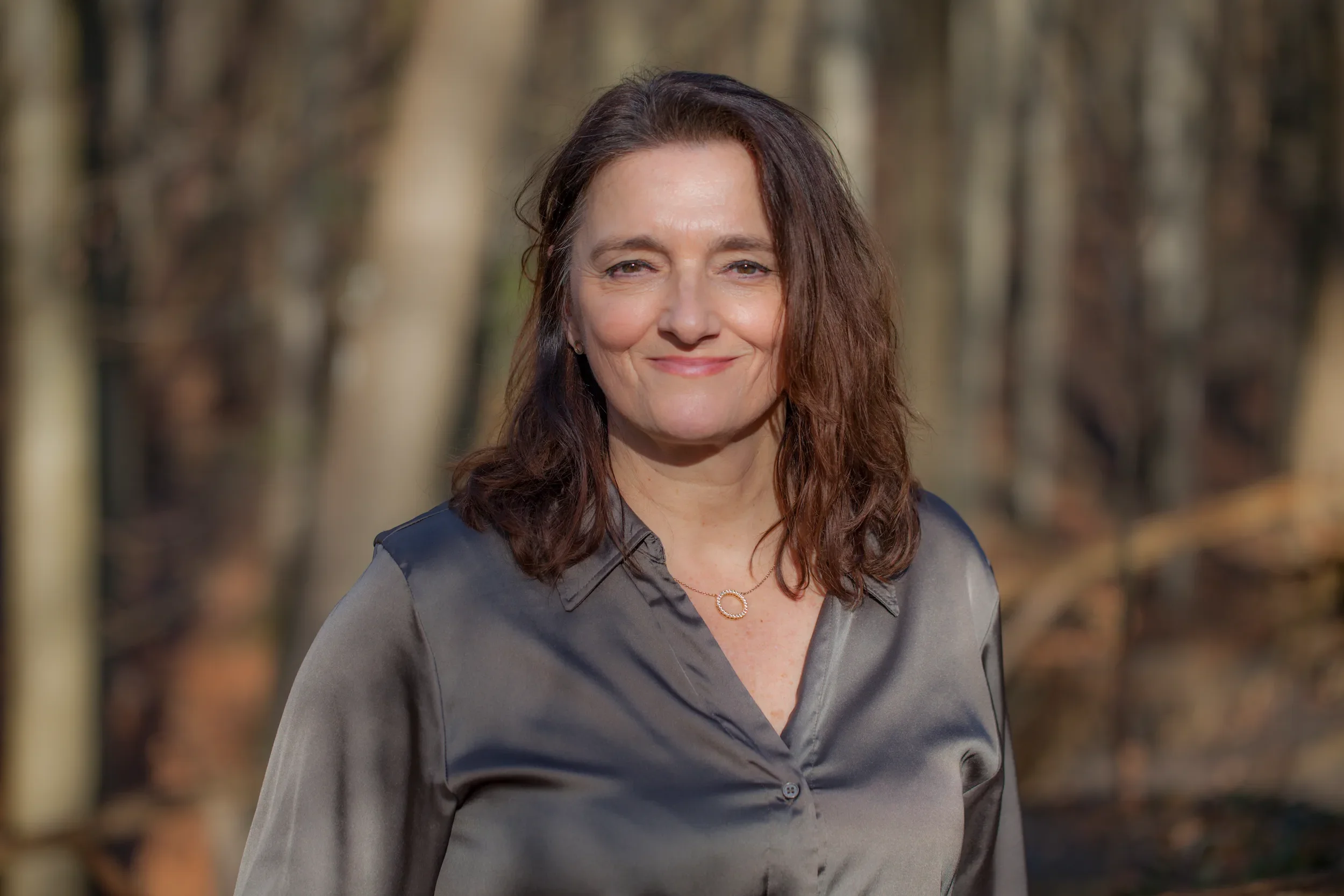 A woman with wavy brown hair wearing a gray button-up shirt and a necklace, standing outdoors in a wooded area with blurred trees in the background.