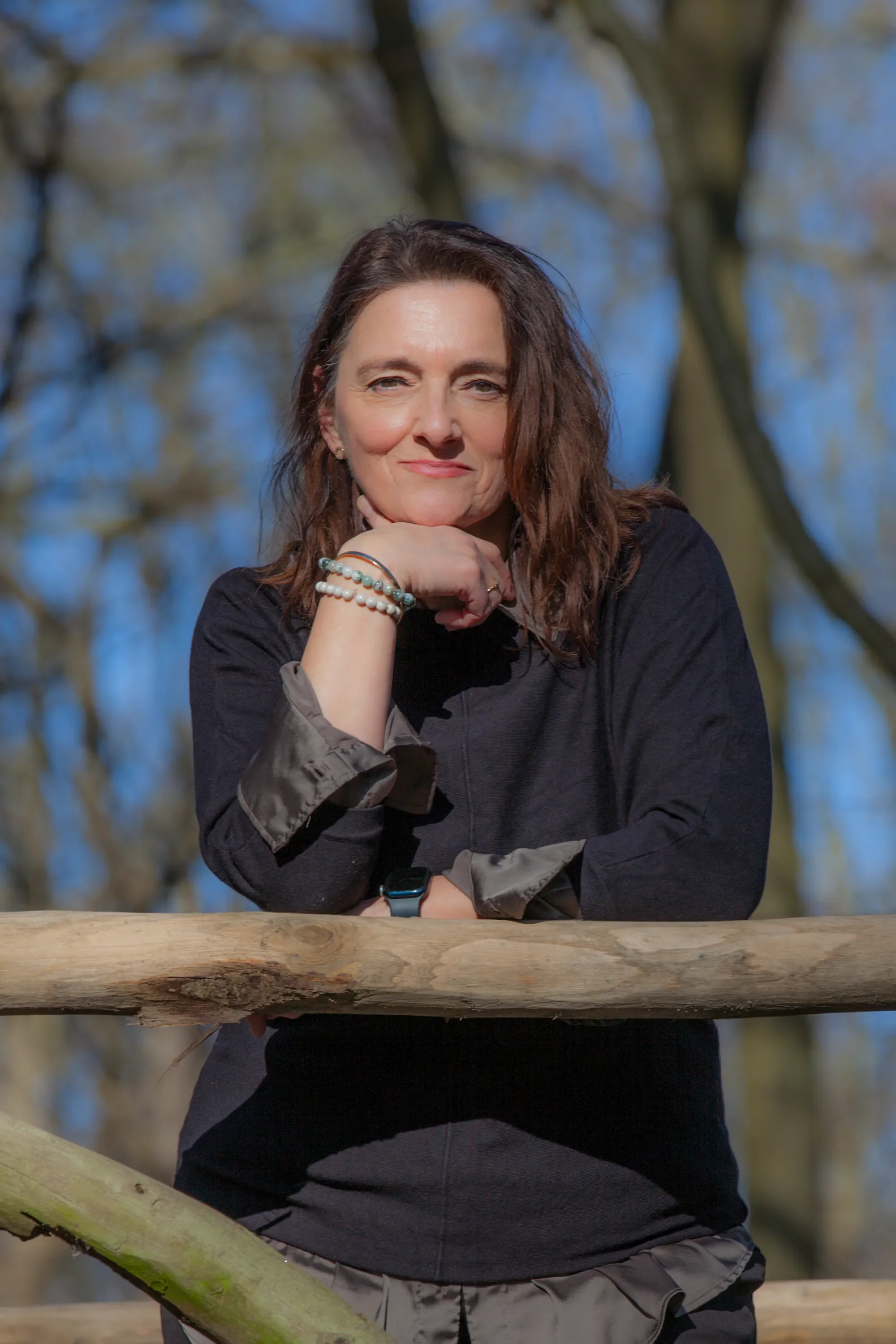 A woman with wavy brown hair standing outdoors, resting her chin on her hand, wearing a black and gray jacket and several bracelets, with a blurred background of trees and blue sky.