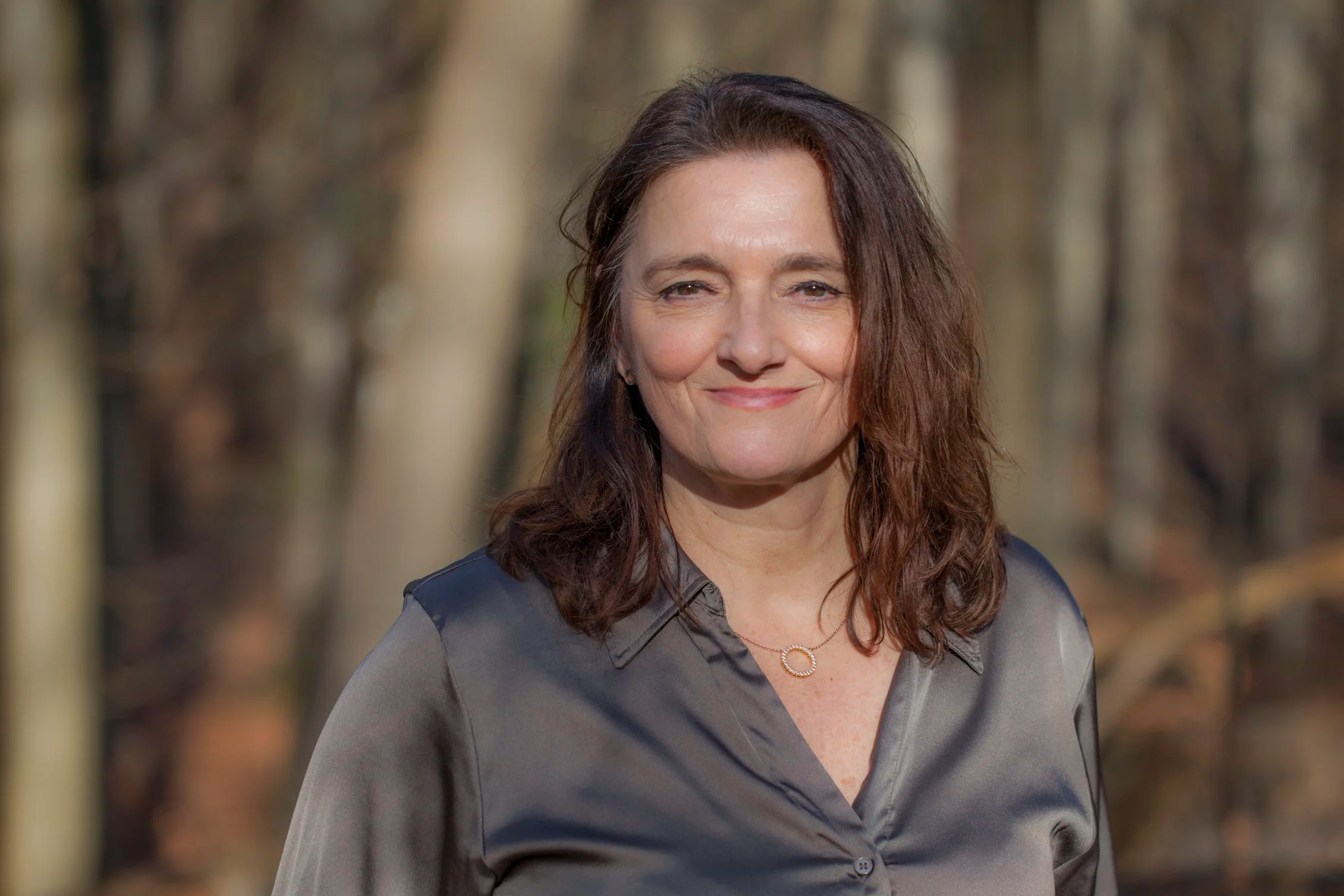 A woman with shoulder-length brown hair, dressed in a gray jacket, smiling outdoors during daytime with a blurred background of trees.