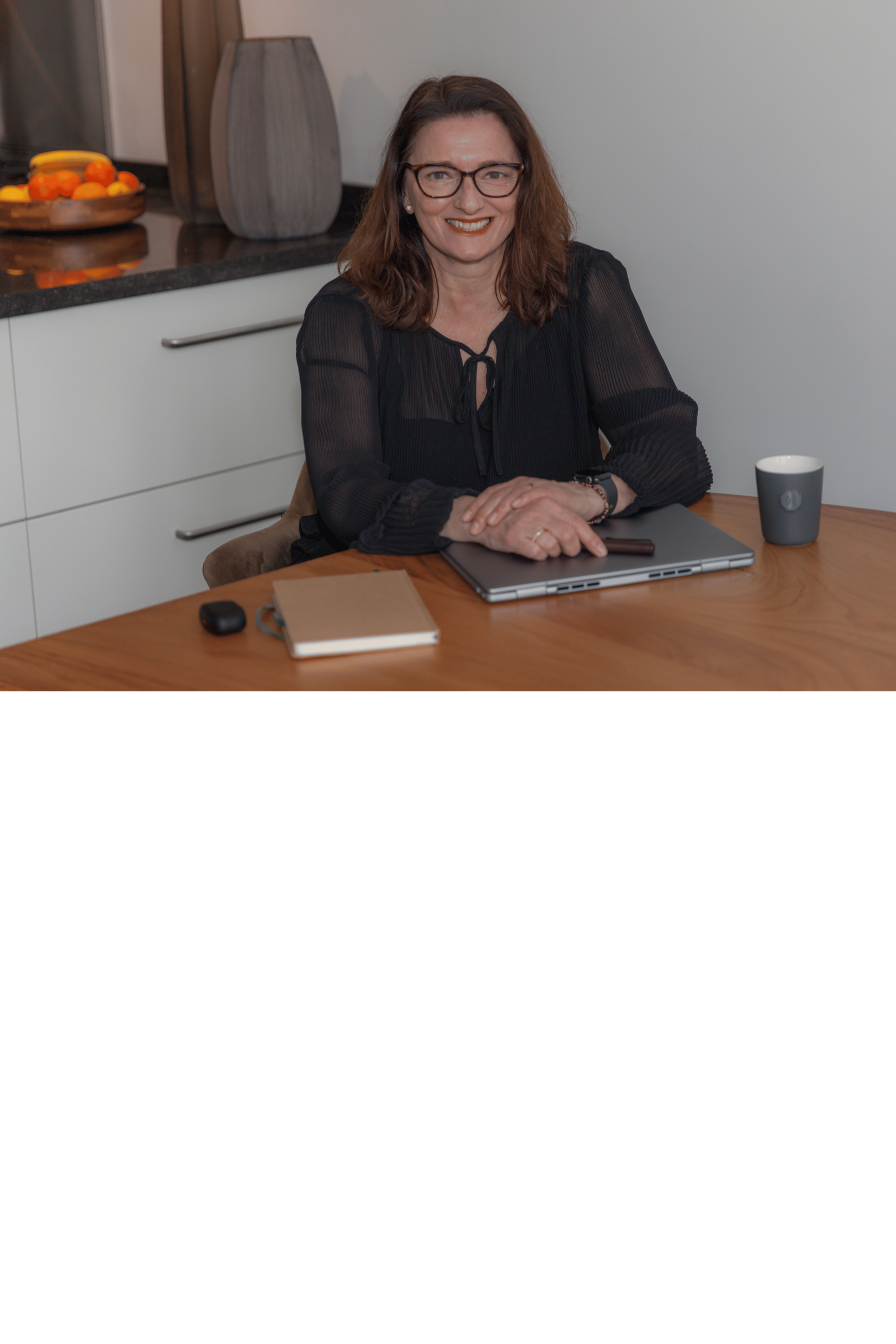A woman with brown hair, glasses, and a black blouse sitting at a wooden table, smiling at the camera. The table has a laptop, a notebook, a computer mouse, a smartphone, and a coffee mug. In the background, there is a kitchen countertop with a bowl of fruit and a decorative vase.