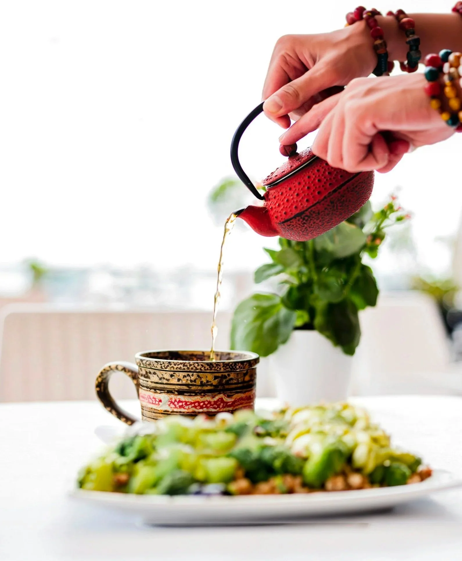 Person watering a potted plant with a red textured teapot-shaped watering can, wearing colorful beaded bracelets, with a plate of salad in the foreground and lush green plants in the background.
