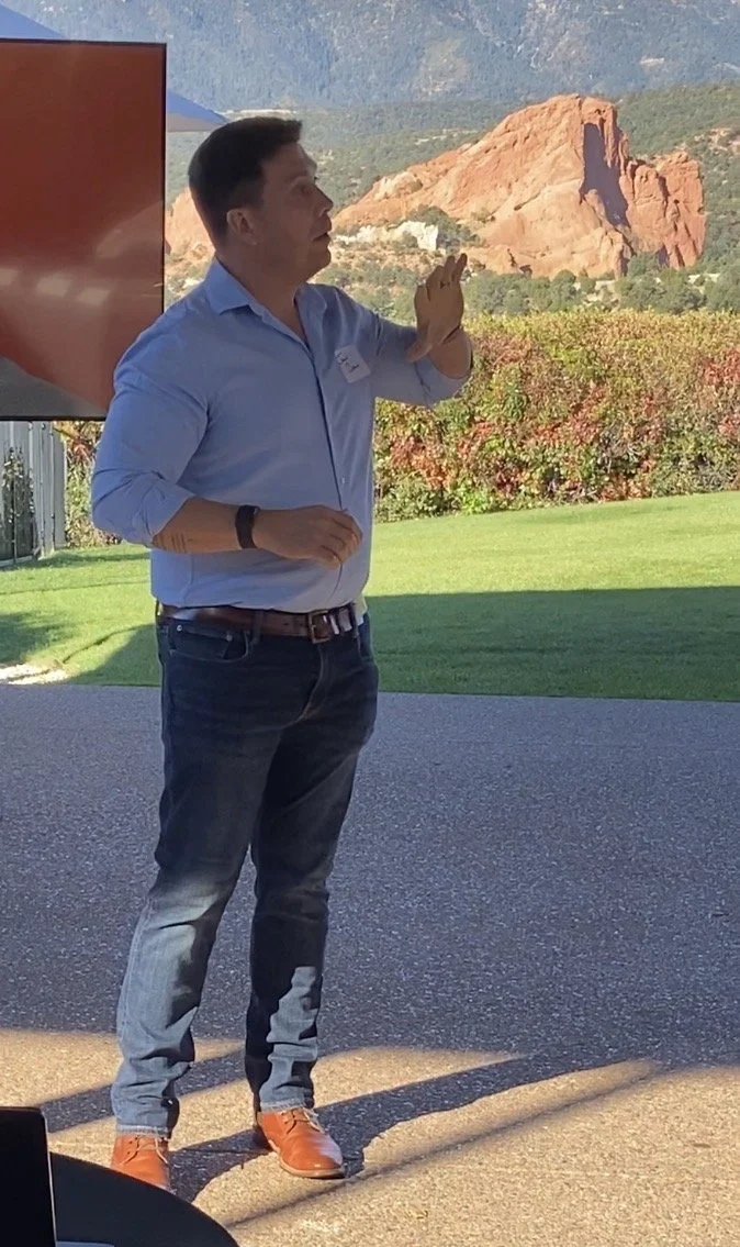 A man in a blue shirt and jeans standing outdoors with mountains in the background, gesturing with his hand during a presentation.