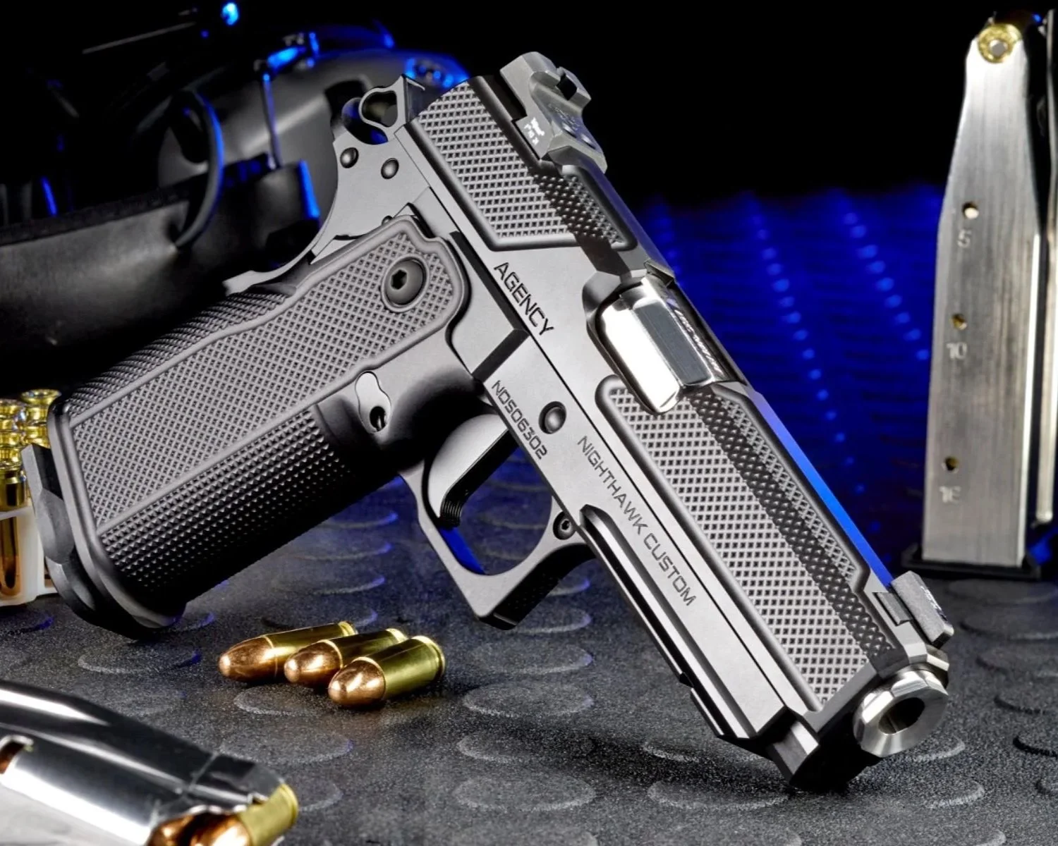 A custom silver handgun with black grips, labeled 'Nighthawk Custom' and 'Agency,' rests on a textured black surface next to several gold and copper-colored bullets, with some firearms magazines in the background.