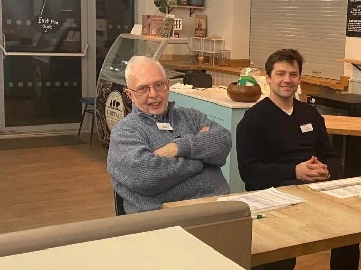 Two men sitting at a wooden table in a cafe, smiling. One older man with white hair and glasses next to a younger man with dark hair. The cafe has a counter with a display case and various items on shelves in the background.