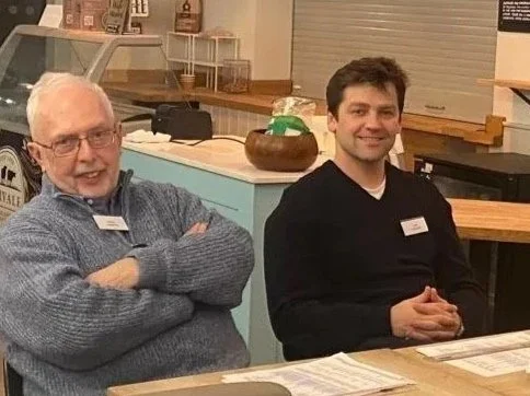 Two men sitting at a table in a casual setting, smiling at the camera.