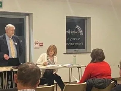 A woman reads or signs documents at a table during a meeting or event, with people seated around her in a room that has a window and a door.
