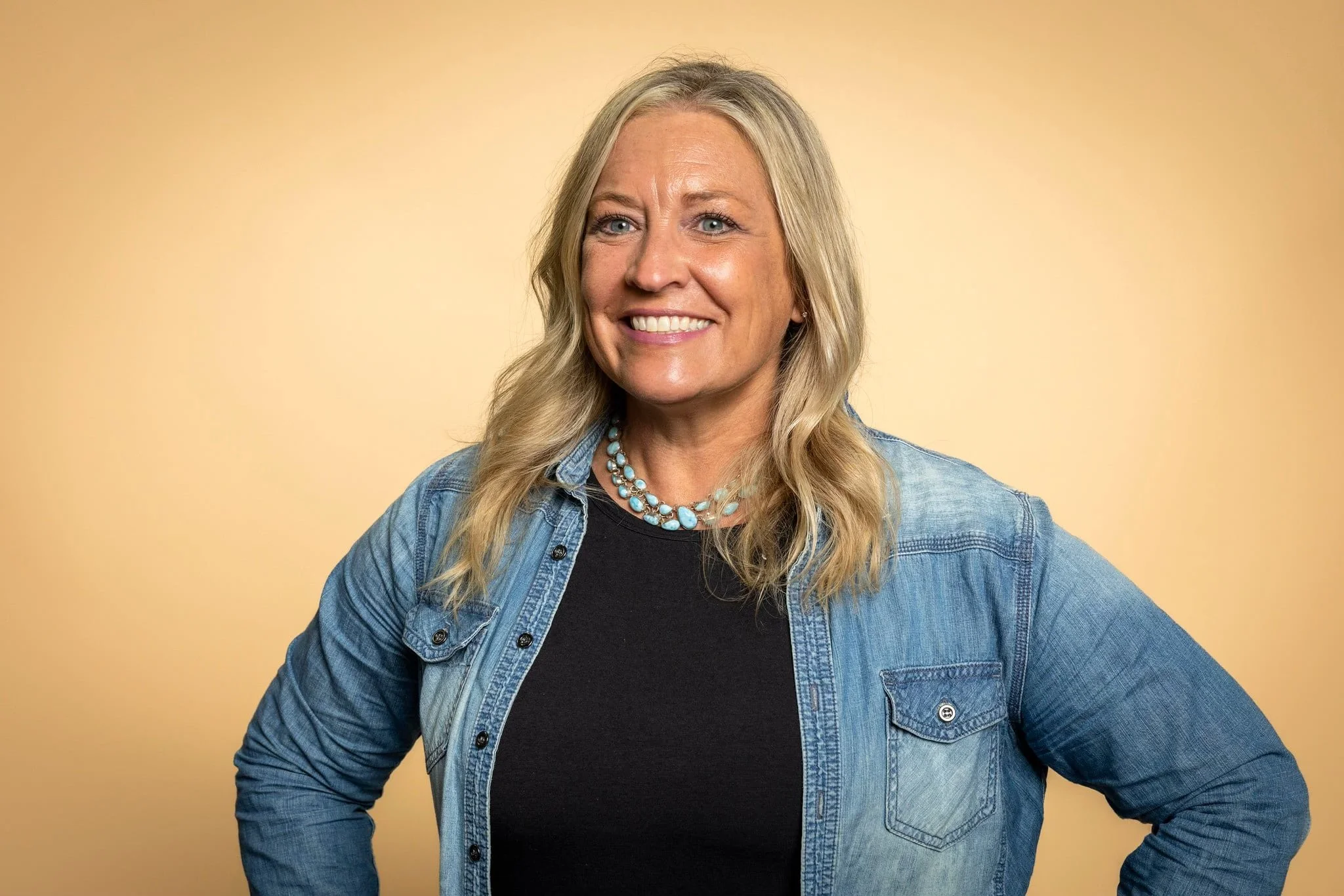 A woman with blonde hair smiling, wearing a denim jacket over a black top and a turquoise necklace, standing with her hands on her hips against a yellow background.