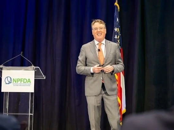 A man in a gray suit, orange tie, and glasses is smiling and holding a microphone in his hands on stage. There is a podium with an NPFDA logo and an American flag behind him. Black curtains are in the background.