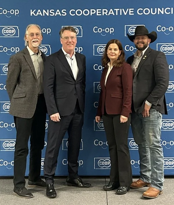 Four individuals standing in front of a blue Kansas Cooperative Council backdrop with Co-op logos, dressed in business and casual attire.
