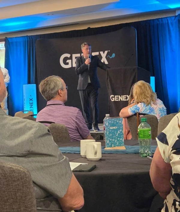 A man in a dark suit speaking at a presentation on a stage with a microphone, behind a black podium with the WORD 'GENEX' on it, in front of an audience seated at tables with various items, including a green bottled drink.