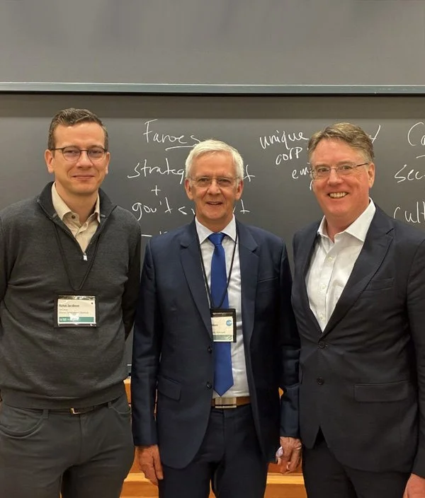 Three men stand together in front of a blackboard in a classroom or conference room, all wearing business attire with name tags. The man on the left is wearing glasses and a sweater, the man in the middle is older with glasses and a suit, and the man on the right is smiling and wearing a suit with an open-collared shirt.