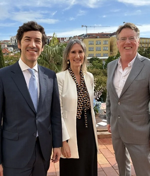 Three people dressed in business attire standing outdoors on a balcony with buildings, trees, and a crane in the background.