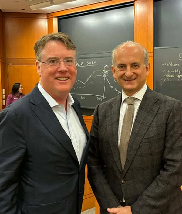 Two men in business suits smiling at a conference or meeting, standing in front of a blackboard with handwritten notes and diagrams.