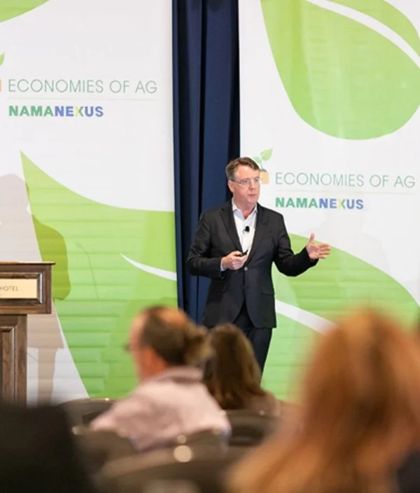 A man in a dark suit giving a presentation at a conference with a background that reads 'Economies of Ag NAMA NEXUS.'