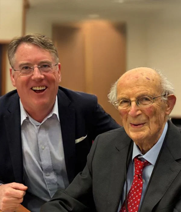 Two men smiling, one younger with glasses and the other elderly with glasses and white hair, both in suits, in a professional setting.