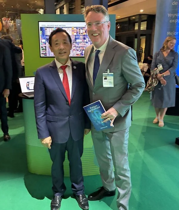 Two men in business suits standing together at a conference or trade show, smiling at the camera. One is holding a booklet titled 'Future Agriculture'. In the background, there is a large digital display, a green booth area, and other people engaging in conversation.