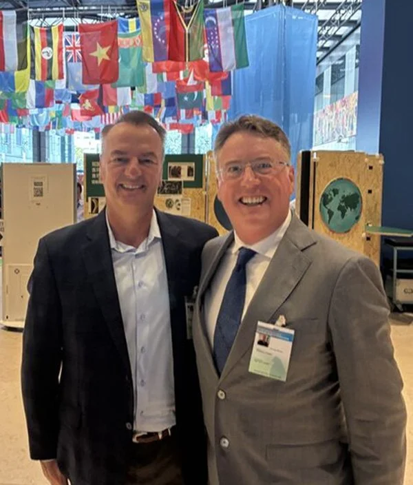 Two men in suits smiling and posing together at an international conference or event, with flags from various countries hanging above them and informational posters displayed behind them.