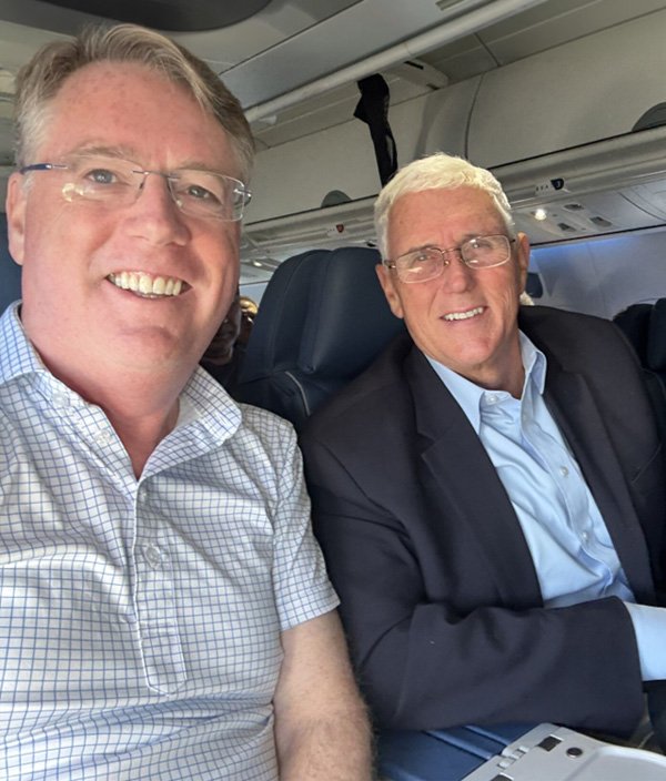 Two men sitting next to each other on an airplane, smiling for a selfie. One has glasses and a checked shirt, the other has glasses and a dark suit.