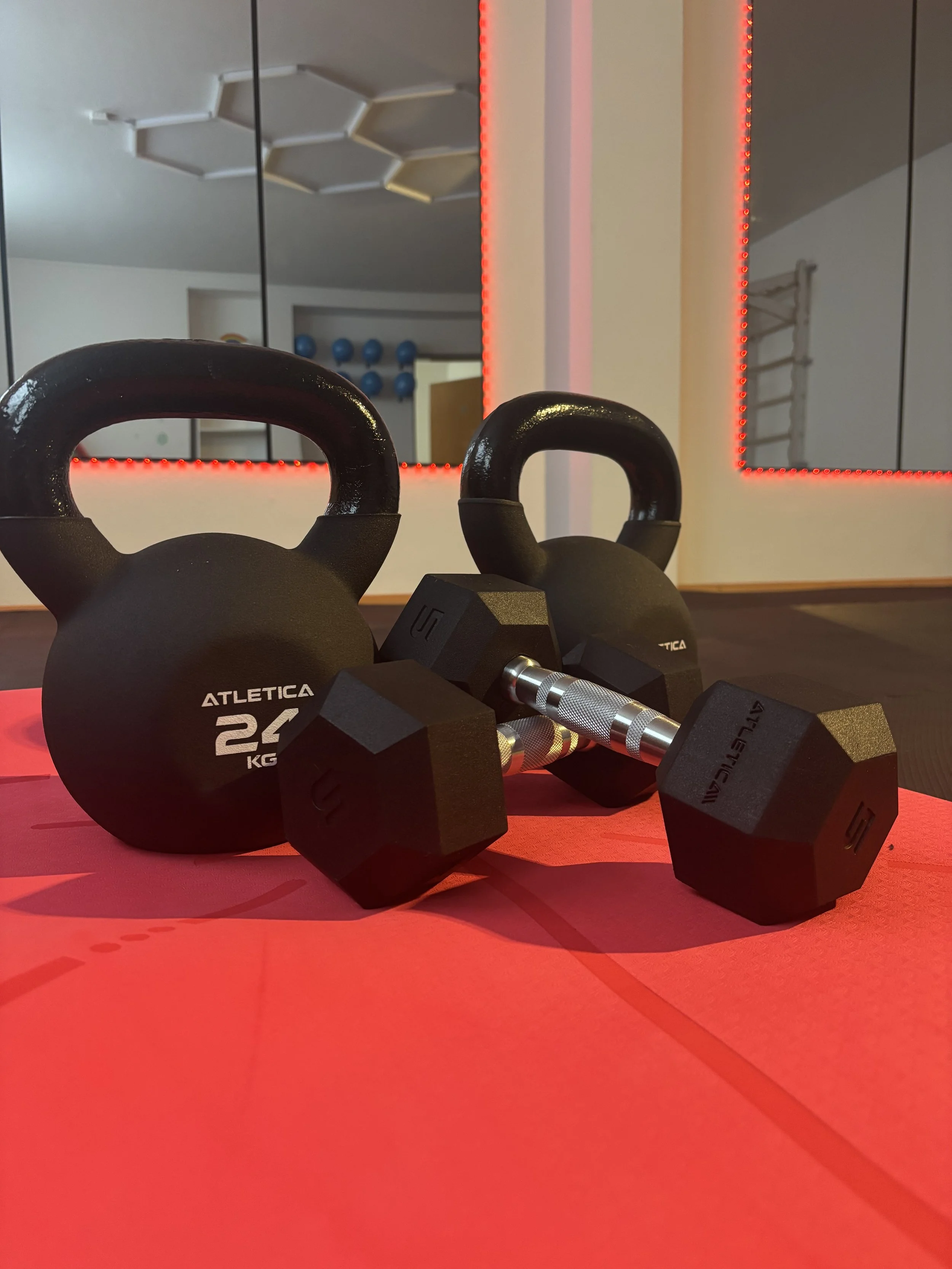 Kettlebells und Hanteln auf rotem Yoga-Mattenboden in einem Fitnessstudio, Spiegel im Hintergrund mit roten Lichtern.