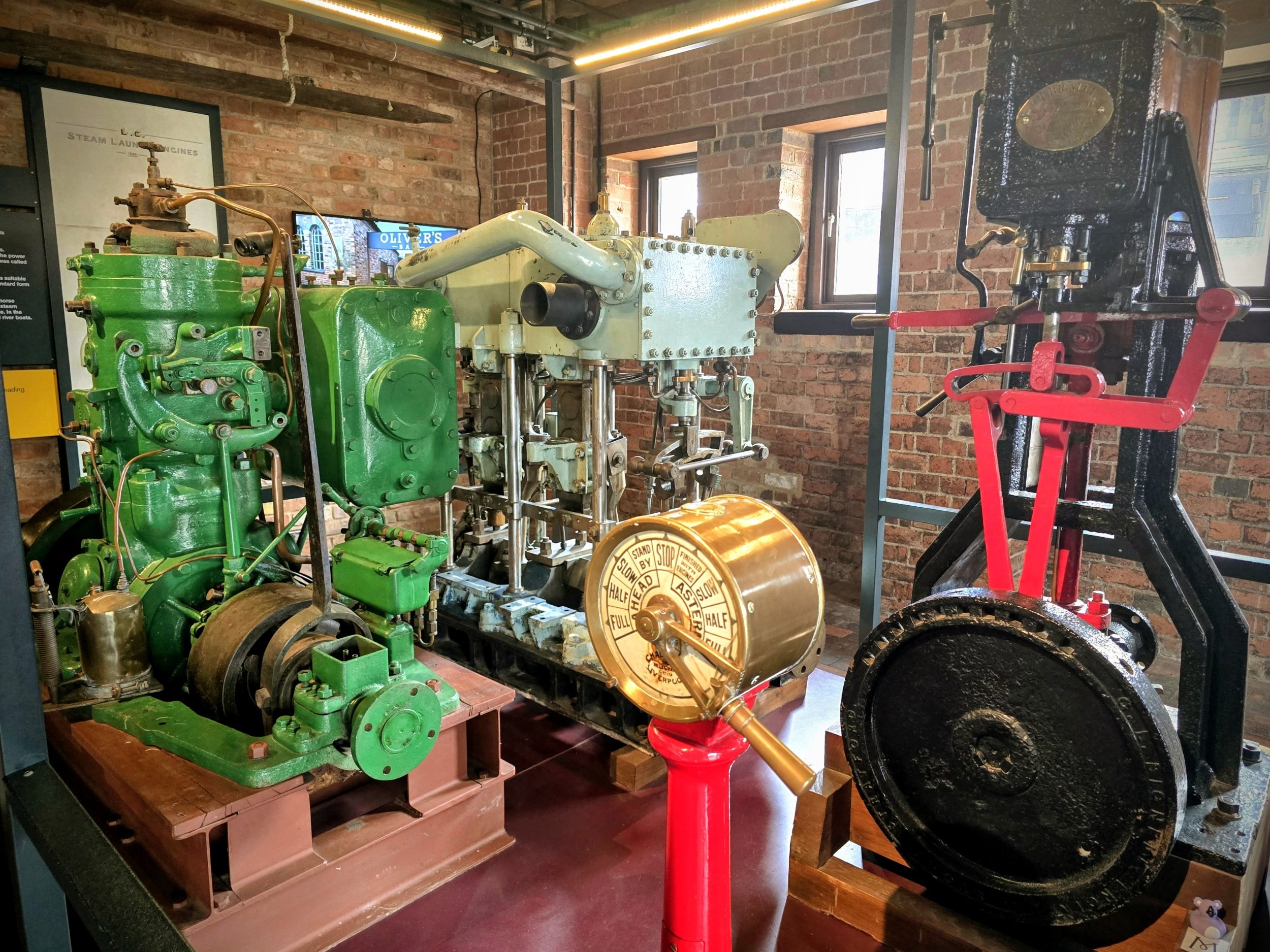 An indoor display of vintage industrial equipment, including a large green machine, a white machine with pipes, and a black and red machine on the right, set against brick walls with windows and a sign.