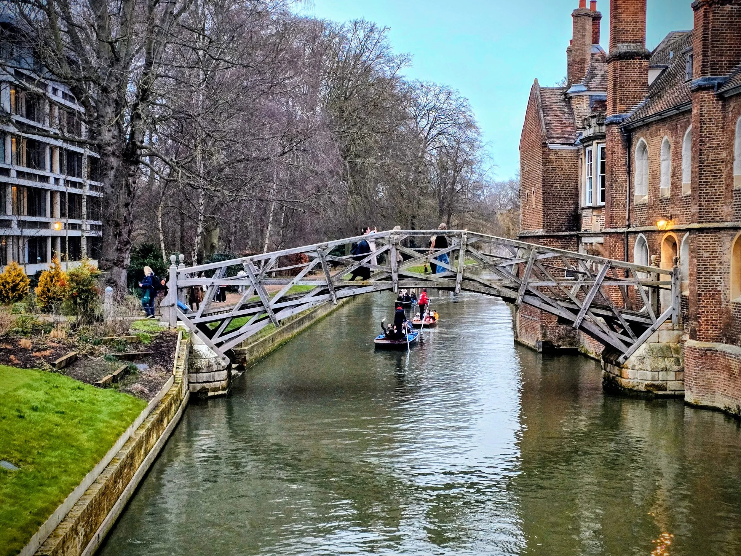 A wooden footbridge over a canal with people paddle-boarding underneath, flanked by trees and brick buildings.