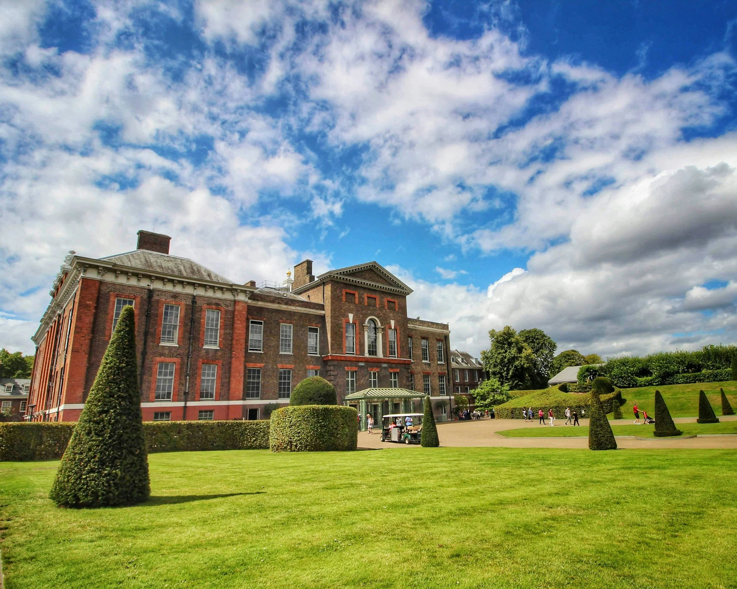 A large historic brick building with white trim and multiple windows, set amidst well-manicured lawns and neatly trimmed topiary, with people walking and a golf cart nearby, under a partly cloudy sky.