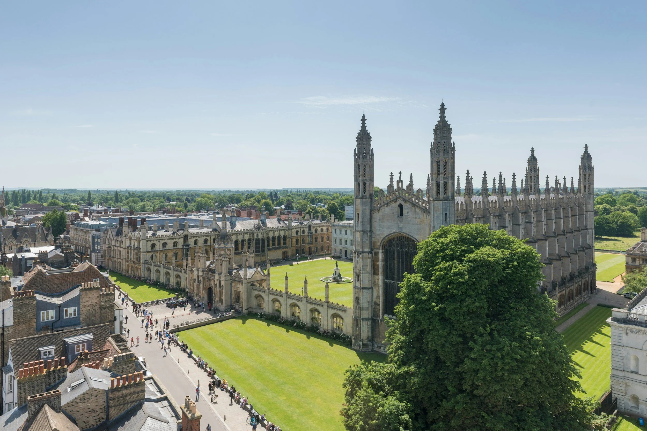 Aerial view of a large historic cathedral with gothic architecture, tall spires, and intricate stone details, surrounded by green lawns and a cityscape./