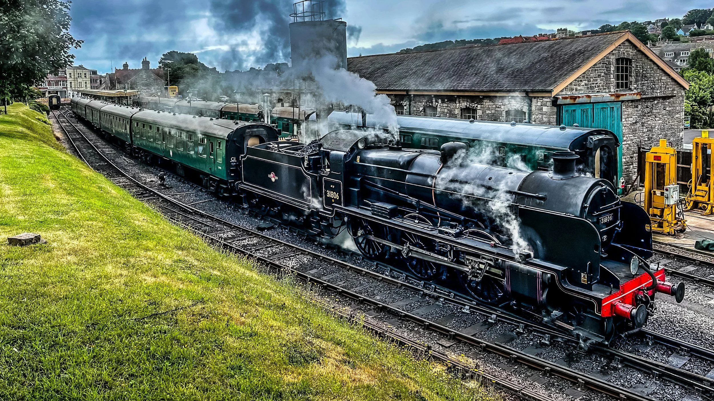 A vintage steam train with black engine and green passenger cars on railway tracks near a stone building, with smoke billowing from the engine.