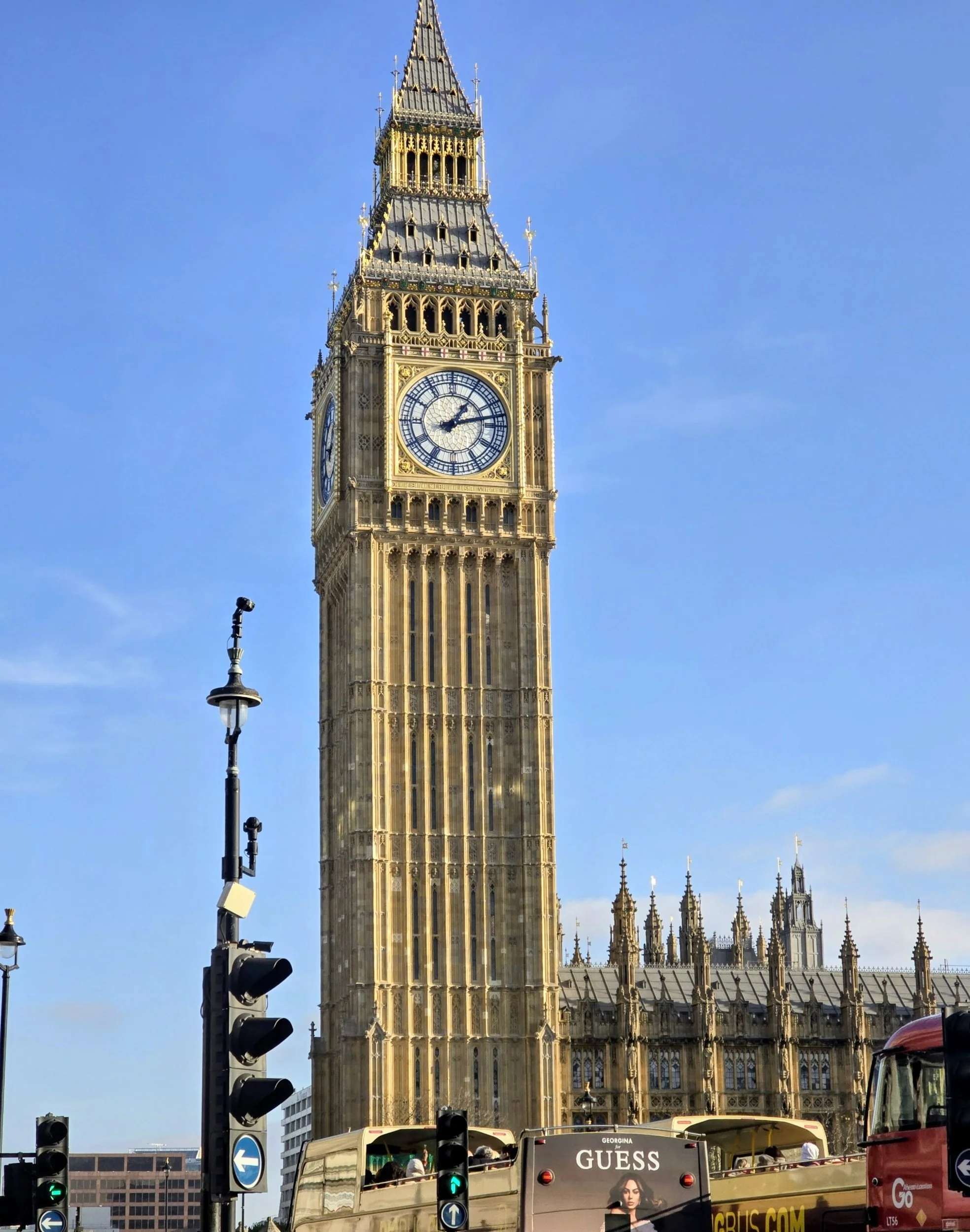 Big Ben clock tower in London, with traffic and buses in the foreground, under a clear blue sky.