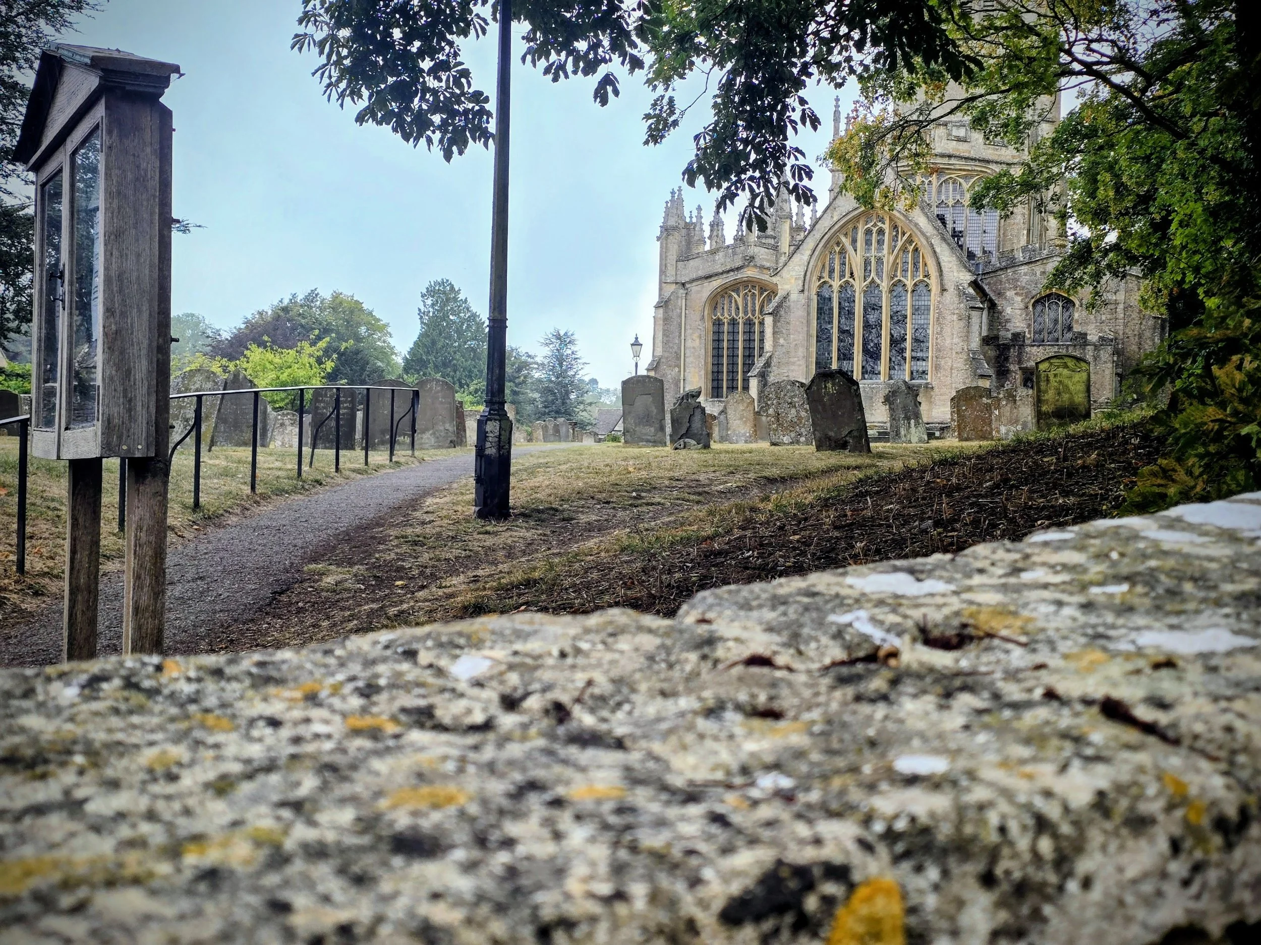 A historic stone church viewed from a low angle with a gravel path, old tombstones, a wooden notice board, and a lamppost in the churchyard, surrounded by trees.