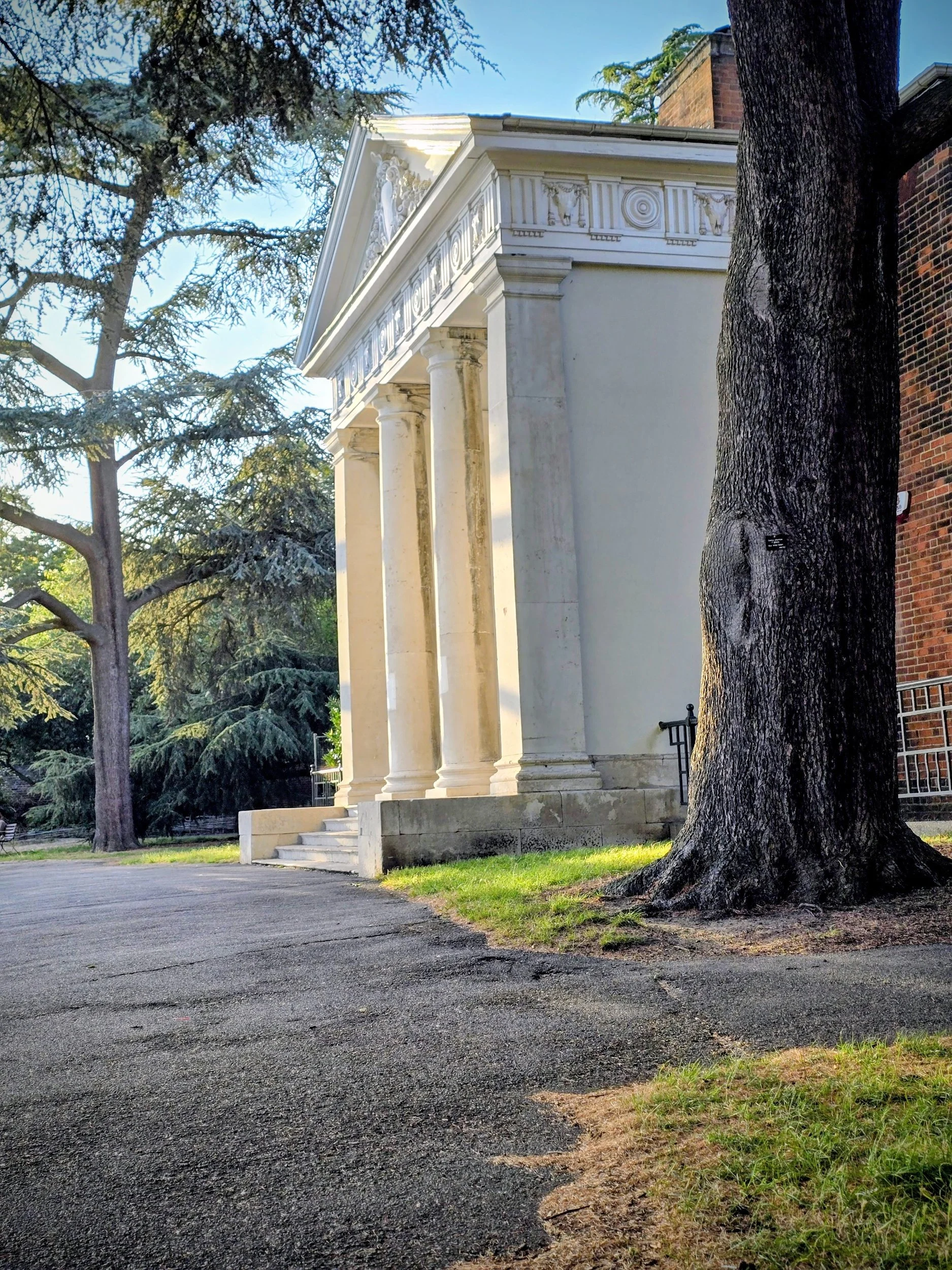 White neoclassical building with columns, stairs, and decorative details, surrounded by trees and greenery.