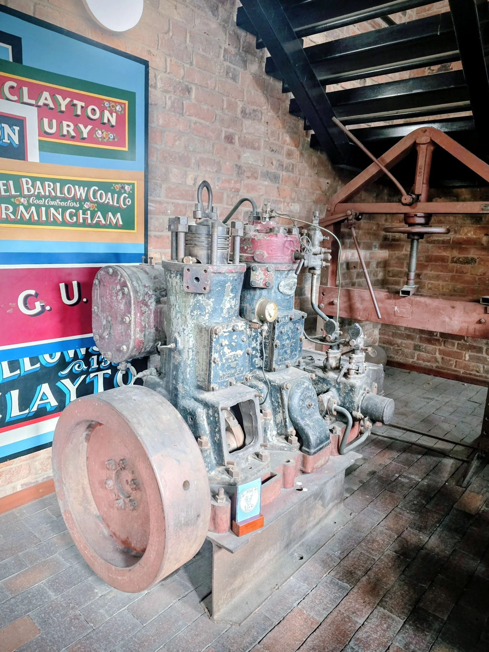 An antique industrial engine on display with a brick wall and colorful sign in the background.